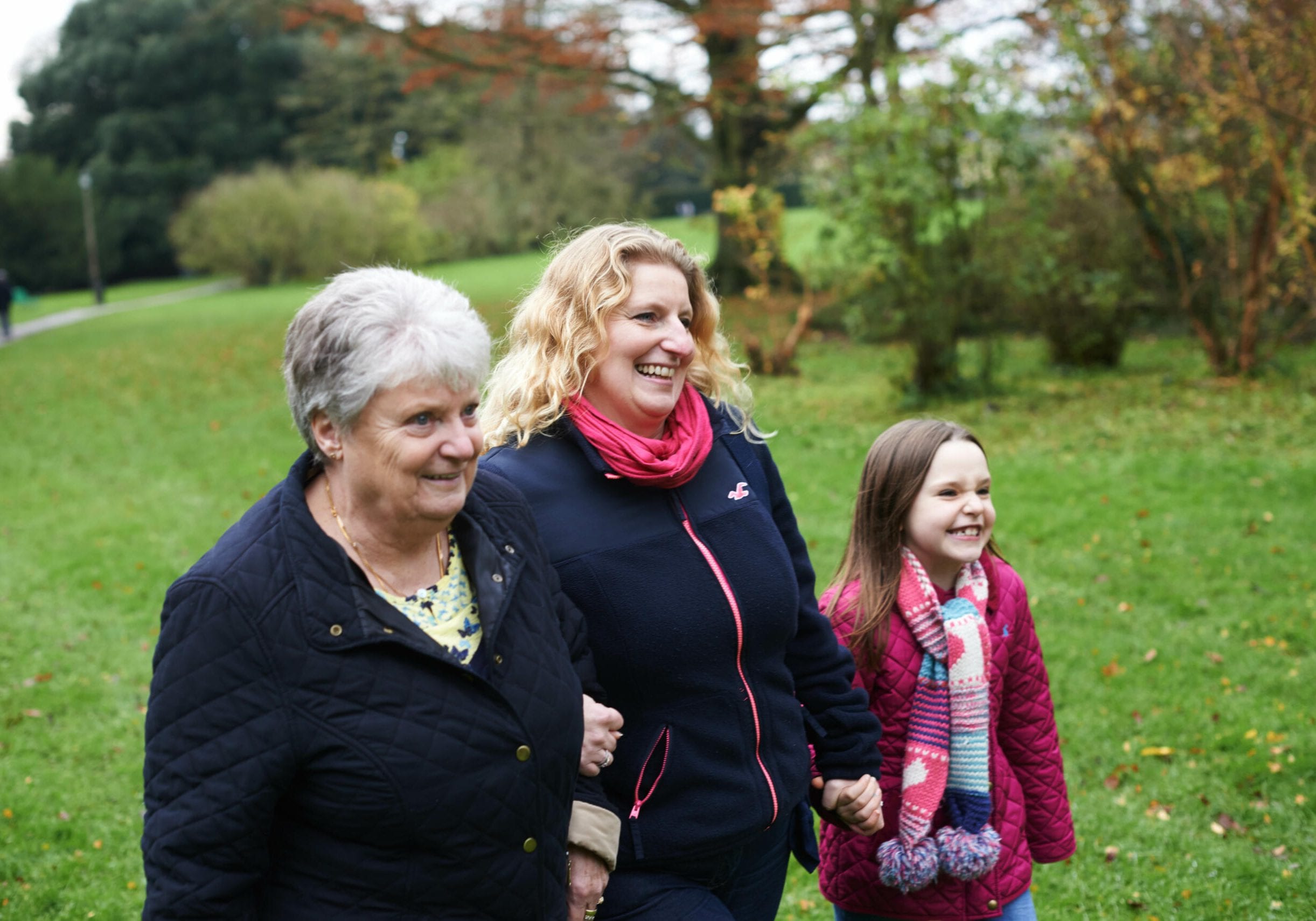 A family walk together in a grassy field, holding hands. There are three women: a young girl, her mother, and her grandmother.